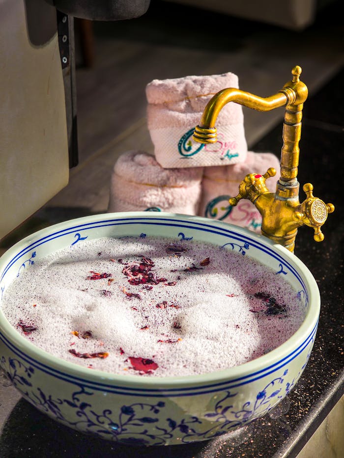 A luxurious spa bowl with rose petals and foam, accompanied by rolled towels and a vintage faucet.